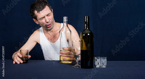 Lonely and drunk man in white home shirt sits at table. In his hands holds glass and bottle of vodka. Drunken look is directed at bottle of alcohol.