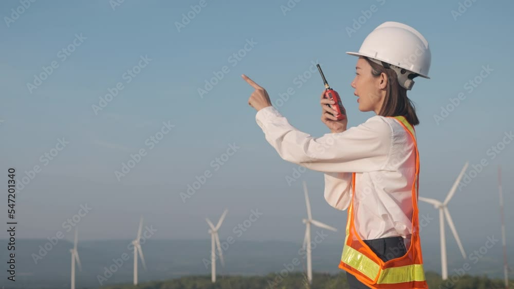 Beautiful asian woman engineer wearing safety helmet and reflective vest pointing finger and talking to her team or someone with radio communication with wind turbine propeller background. Beautiful asian woman engineer wearing safety helmet and reflective vest pointing finger and talking to her team or someone with radio communication with wind turbine propeller background.