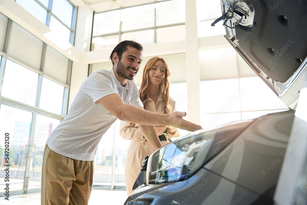 Pleased young man inspecting new motorcar in presence of saleswoman Pleased young man inspecting new motorcar in presence of saleswoman