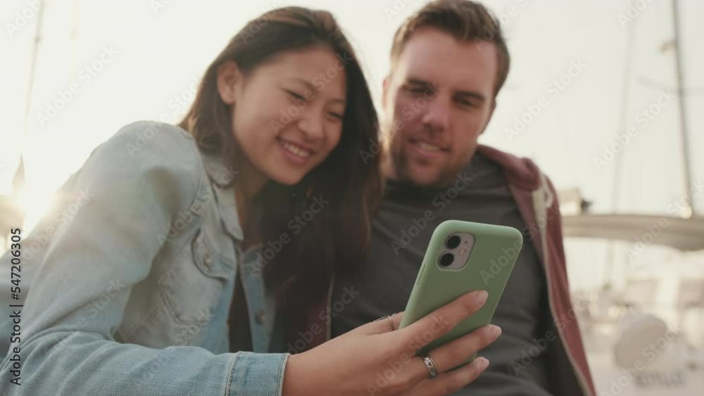 Happy couple sitting in the harbor and watching photos, videos on mobile phone Happy couple sitting in the harbor and watching photos, videos on mobile phone