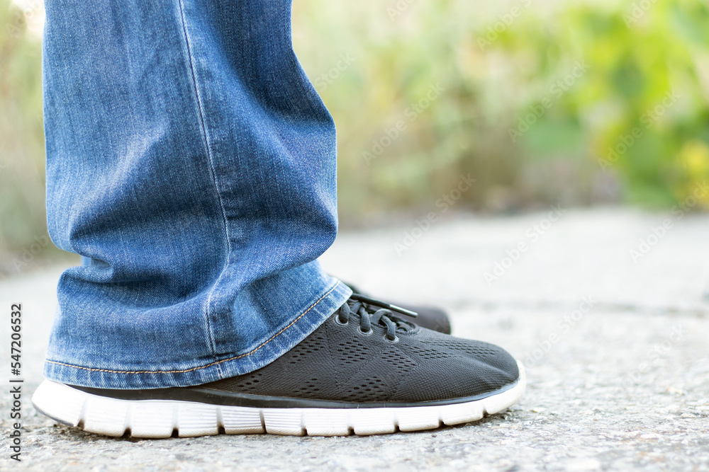 A man in blue jeans and black sneakers stands on a concrete road. Close-up with a blurred background.