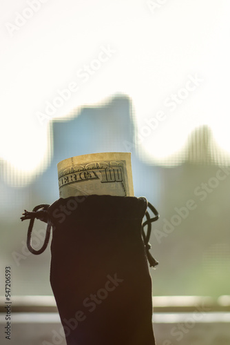 A hundred dollar bill sticks out of a black velvet pouch in bright sunlight. Close-up with a blurred background.