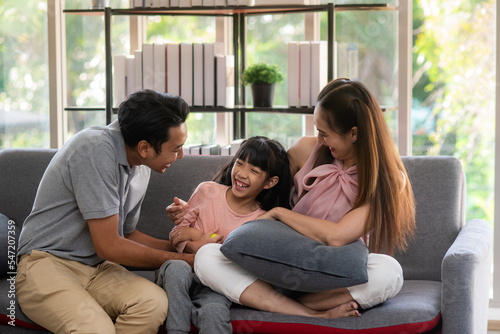 Happy asian family, young father, mother and adorable daughter on sofa inside living room Tease and tickle each other Laughing happily, looking warm close relationship.