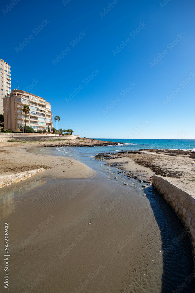 El agua del río acabando en el mar Mediterráneo de la playa de Calpe en Alicante bajo un cielo azul con varios hoteles de fondo.