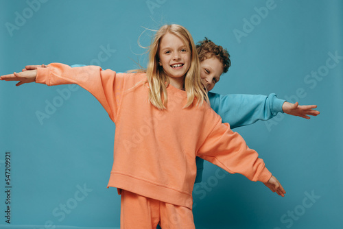 cute, beautiful, happy children a boy and a girl of school age stand on a blue background in bright plain clothes, facing the camera spreading their arms to the sides