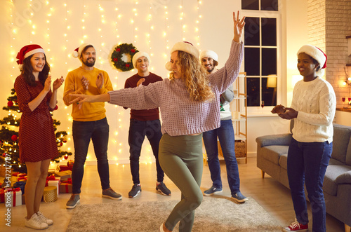 Happy young diverse people in Santa hats having fun at Christmas party at home. Group of cheerful millennial multi ethnic friends applauding dancing woman in Santa cap and trendy crop top sweater