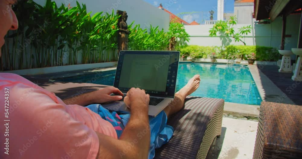 Rear view of man blogger remote working on laptop during vacation near swimming pool. Guy uses notebook typing on computer lying os a sun lounger.