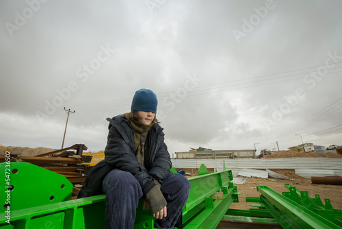 cool stylish caucasian boy with the blue hat on his face covering his eyes in a coat and hat with a scarf posingon green metal structures in an industrial area against a cloudy greysky on a rainy day photo