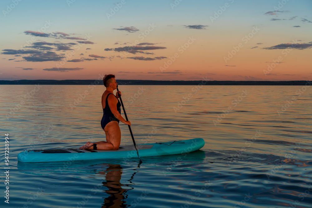 a Jewish feminist woman in a closed bathing suit with a mohawk on a koyen on a stand-up board with an oar floats on the water against the background of the sunset sky.