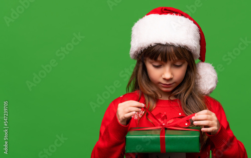 A cute little girl in a Santa hat unpacks a New Year's gift. Merry Christmas and Happy New Year 2023. Portrait isolated on green background