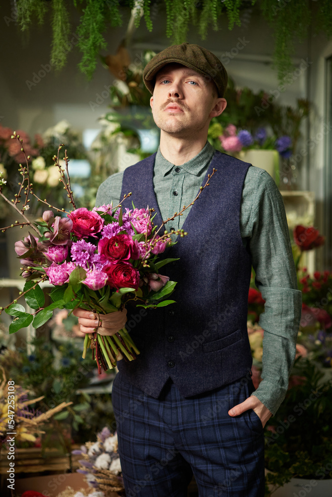 Fototapeta premium Attractive cheerful caucasian male in peaked cap and vintage clothes from 20s holding beautiful bouquet of red and violet flowers. Mother's Day or Valentine's Day concept. High quality image