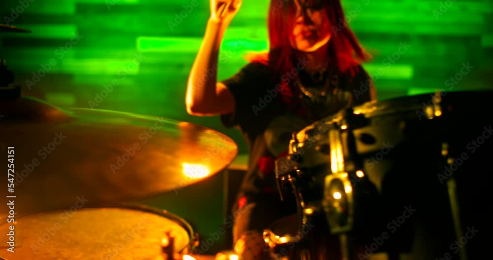 Drummer girl vigorously playing drums in green and orange flickering strobe lights on the nightclub stage. Performance of female rock band. Focus shifts from the drum set to the girl and back again. Drummer girl vigorously playing drums in green and orange flickering strobe lights on the nightclub stage. Performance of female rock band. Focus shifts from the drum set to the girl and back again.