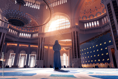Fotografia, Obraz Back view modern woman in traditional blue abaya standing in Grand Mosque with magic sun light