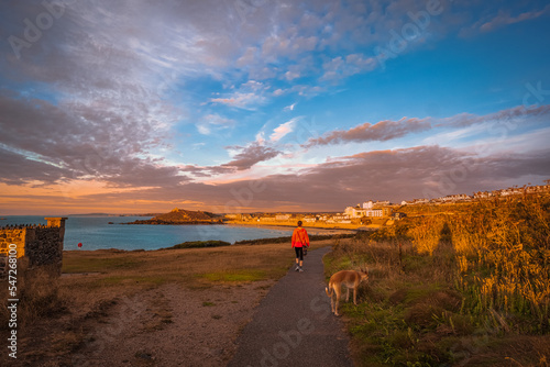 Beautiful view of rugged coastline at sunset in summer with silhouette of walking woman; woman's dog in foreground and seaside town and dramatic sky in background