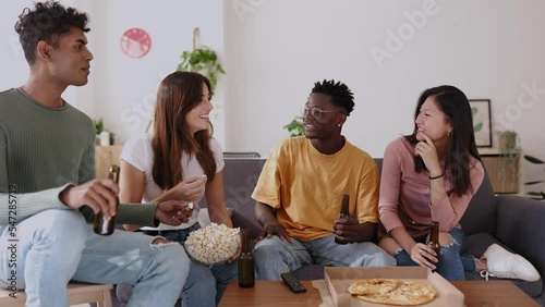 Happy young friends sitting on sofa watching TV together at home. Young group of teenage people having fun together social gathering eating pizza and drinking beer in the living room.  photo