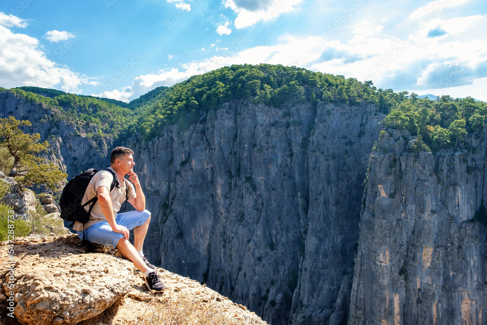 Naklejka premium Side view of tranquil male hiker with backpack sitting on edge of rocky cliff in highlands on summer day