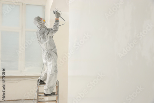 Decorator in protective overalls painting wall with spray gun indoors © New Africa