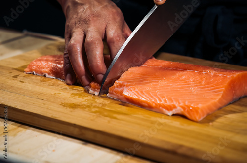 Close up of Chef cook hands chopping salmon fish for traditional Asian cuisine with Japanese knife. Professional Sushi chef cutting seafood japanese chefs are making salmon fish sashimi. dark tone