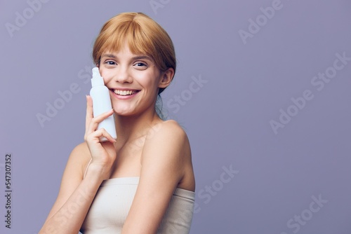 A gorgeous, joyful woman in a white T-shirt with fresh, well-groomed skin, with red wavy hair gathered in a tail stands on a lilac background with spray.Horizontal studio shot.
