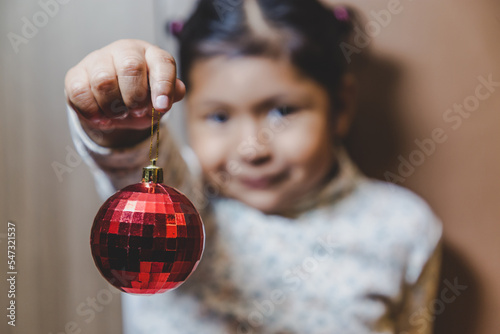 Obraz na plátně niña feliz y sonriente sosteniendo en sus manos objetos decorativos navideños, bolas o esferas navideñas  y letras de feliz navidad en ingles, concepto navidad, amor, paz y compartir