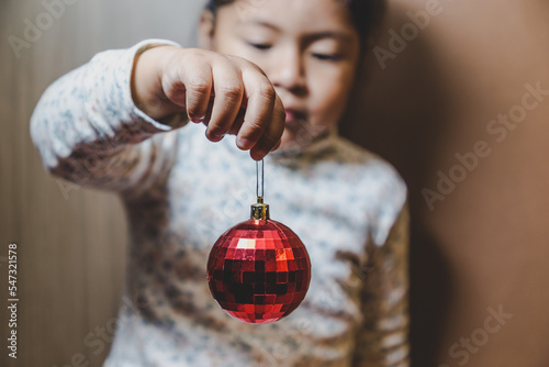Fotografie, Obraz niña feliz y sonriente sosteniendo en sus manos objetos decorativos navideños, bolas o esferas navideñas  y letras de feliz navidad en ingles, concepto navidad, amor, paz y compartir