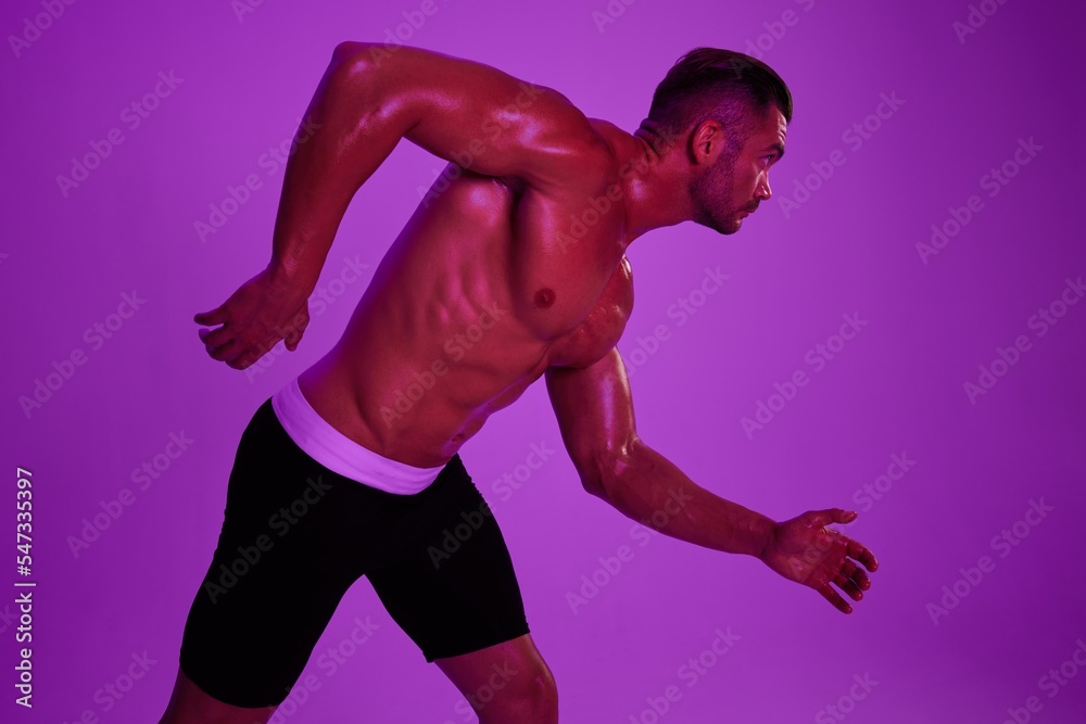 Man athletic body bodybuilder in briefs with nude torso abs full-length in the background fitness classes, purple colored light. Advertising, sports, active lifestyle, competition, challenge concept. Man athletic body bodybuilder in briefs with nude torso abs full-length in the background fitness classes, purple colored light. Advertising, sports, active lifestyle, competition, challenge concept.
