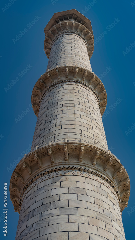 The minaret of the ancient Taj Mahal against the blue sky. A tall white marble tower with carved balconies. India. Agra