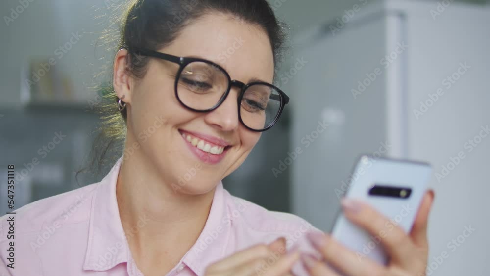 Cheerful positive girl in stylish eyeglasses typing on smartphone, leaving comments to posts of her friends or favourite bloggers, admiring news feed in social networks, digitalization Cheerful positive girl in stylish eyeglasses typing on smartphone, leaving comments to posts of her friends or favourite bloggers, admiring news feed in social networks, digitalization