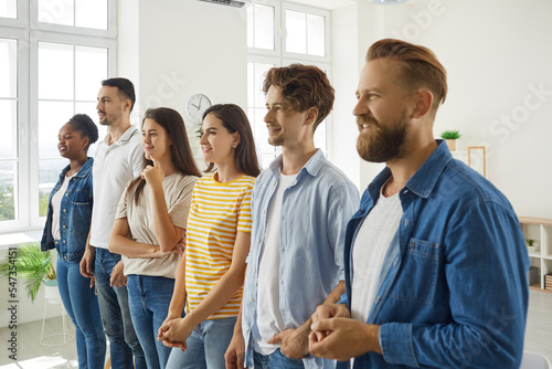 Diverse young people in casual clothes listen to their coach during personal development training. Smiling multiracial men and women stand in row in bright office and listen attentively to someone.