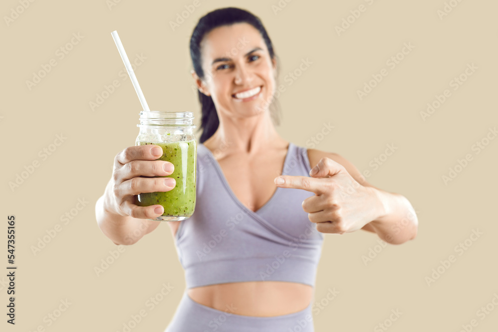 Caucasian woman holds out appetizing green smoothie in jar with straw, offering you to try healthy food from fresh vegetables to maintain vegetarian diet, stands in beige studio. Selective focus Caucasian woman holds out appetizing green smoothie in jar with straw, offering you to try healthy food from fresh vegetables to maintain vegetarian diet, stands in beige studio. Selective focus