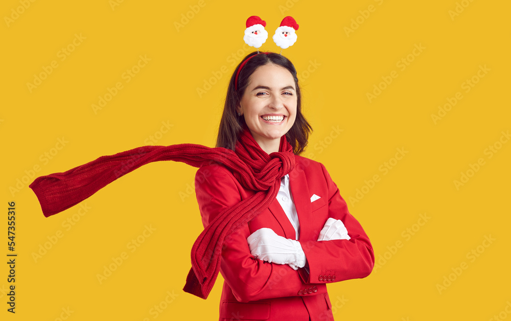 Beautiful brunette is smiling sweetly, dressed in a red business suit with Santa Claus figurines on her head and a red scarf. The model stands with her arms crossed on an isolated yellow background. Beautiful brunette is smiling sweetly, dressed in a red business suit with Santa Claus figurines on her head and a red scarf. The model stands with her arms crossed on an isolated yellow background.