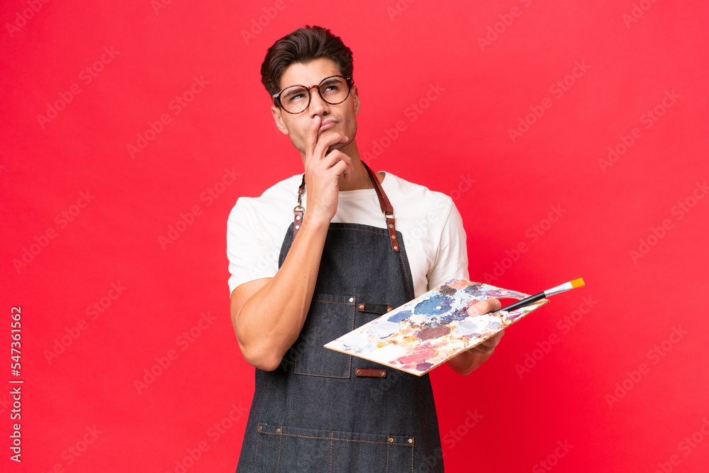 Young artist caucasian man holding a palette isolated on red background having doubts while looking up Young artist caucasian man holding a palette isolated on red background having doubts while looking up