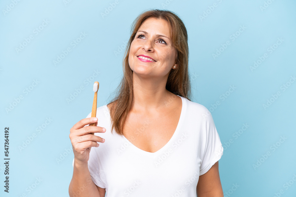 Middle-aged caucasian woman brushing teeth isolated on blue background looking up while smiling Middle-aged caucasian woman brushing teeth isolated on blue background looking up while smiling