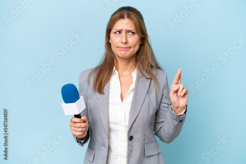 Middle-aged TV presenter woman over isolated blue background with fingers crossing and wishing the best