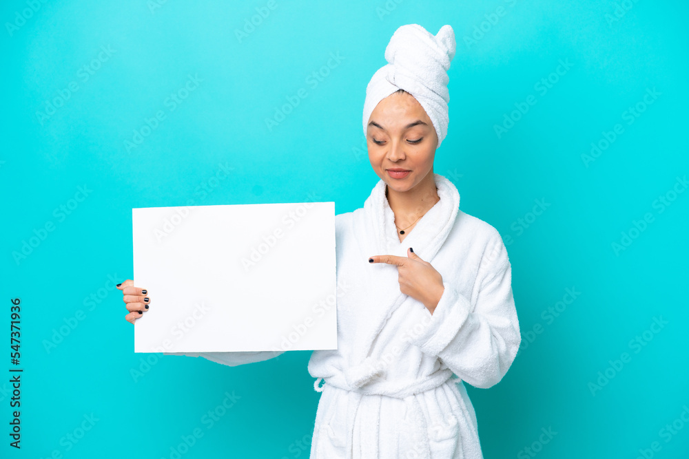 Young woman in a bathrobe with towel isolated on blue background holding an empty placard with happy expression and pointing it Young woman in a bathrobe with towel isolated on blue background holding an empty placard with happy expression and pointing it