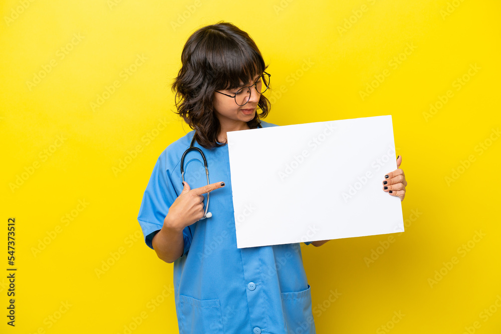 Young nurse doctor woman isolated on yellow background holding an empty placard with happy expression and pointing it Young nurse doctor woman isolated on yellow background holding an empty placard with happy expression and pointing it