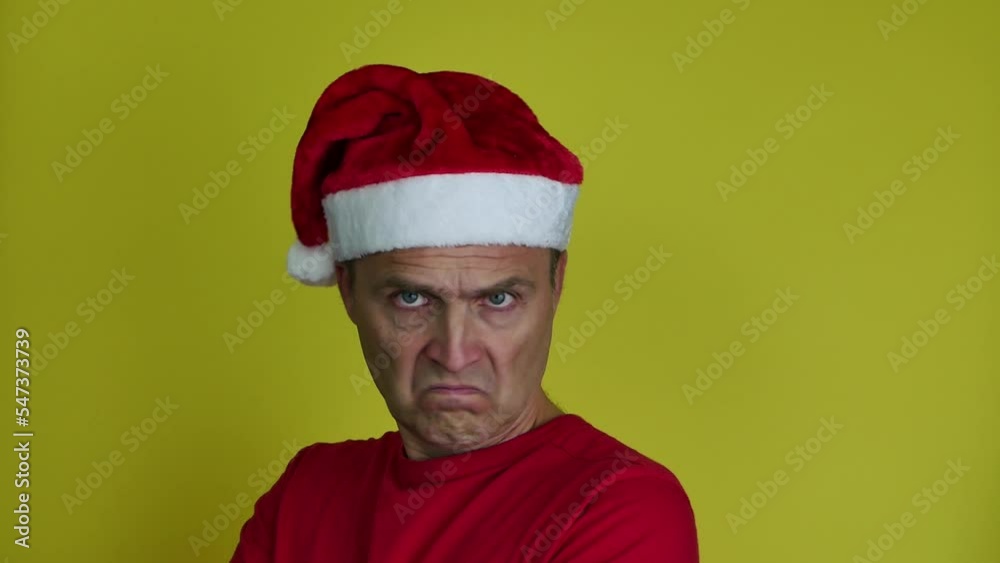 A handsome man in a Santa hat in a red T-shirt on a yellow background is angry and looks sullenly at the camera. A man shows negative emotions. Bad vacation. Bad Christmas. A handsome man in a Santa hat in a red T-shirt on a yellow background is angry and looks sullenly at the camera. A man shows negative emotions. Bad vacation. Bad Christmas.