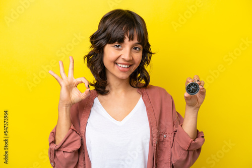 Young latin woman holding compass isolated on yellow background showing ok sign with fingers