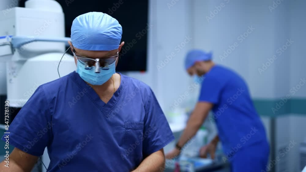 Neurosurgeon in device glasses and mask standing in the surgery room. Doctor is speaking and gesturing with hands. Medical staff at backdrop. Neurosurgeon in device glasses and mask standing in the surgery room. Doctor is speaking and gesturing with hands. Medical staff at backdrop.