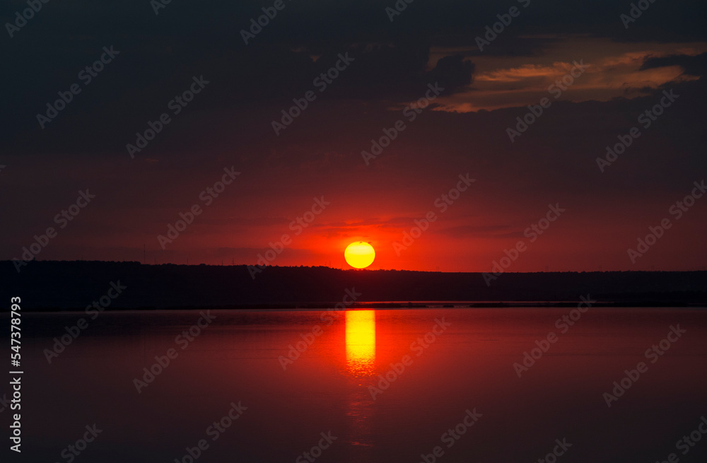A large round sun sets on the horizon and is reflected in the water of the estuary