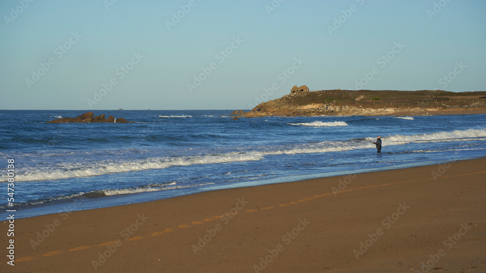 Fisherman with fishing rod on the beach. Rocks in blue sea and hills landscape. Grey sky for copy space. France, Brittany, Kerhillio.