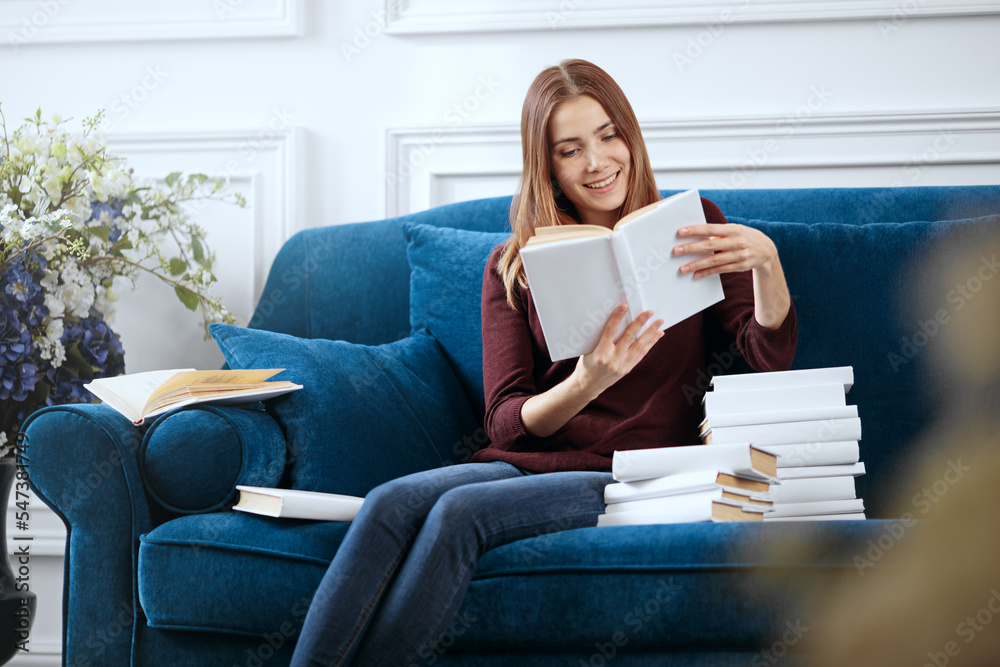 Young smiling woman is reading on a sofa with a pile of books.