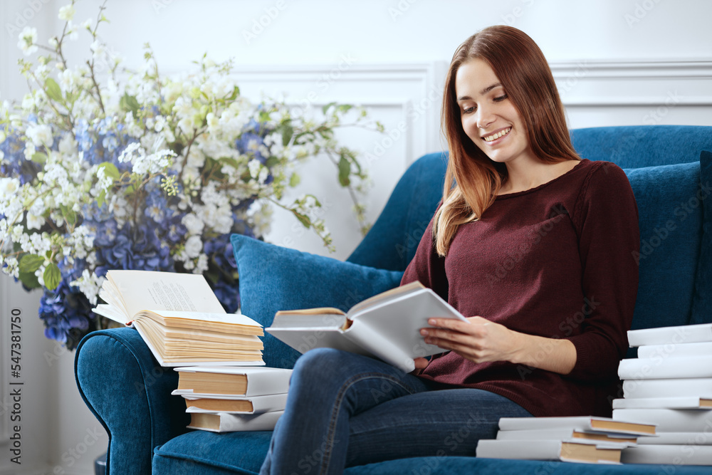 Young smiling woman among a pile of books is reading on a sofa.