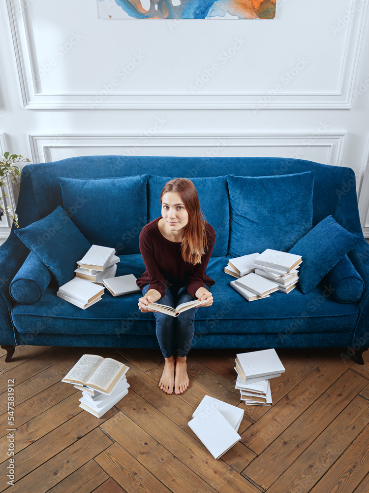 Young surprised woman among stacks of books on a sofa in the living room.