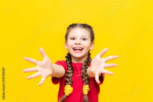 The little girl pulls her hands forward showing her palms and smiles. A beautiful child with pigtails in a red T-shirt on a yellow isolated background.