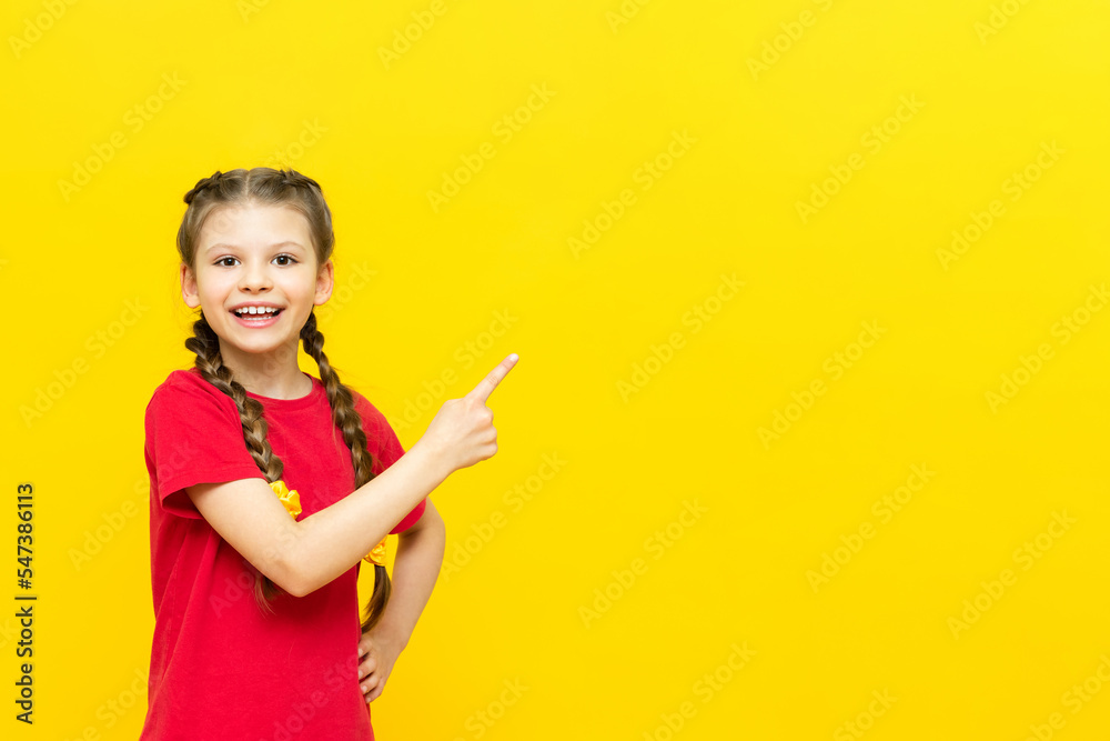 A little girl points her finger at an empty space for your advertisement on a yellow isolated background. A beautiful child is smiling in a red T-shirt with two pigtails. Copy space. A little girl points her finger at an empty space for your advertisement on a yellow isolated background. A beautiful child is smiling in a red T-shirt with two pigtails. Copy space.