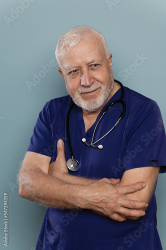 Elderly man doctor standing with stethoscope in medical clothes on blue background, family medicine concept