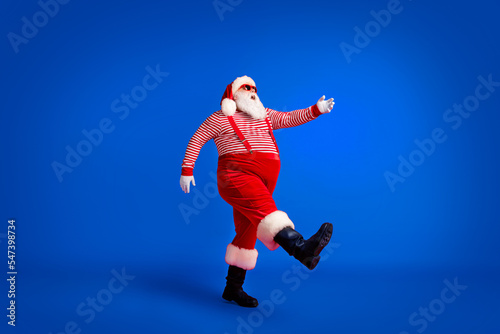 Full length profile photo of grandfather grey beard step raise hand wear santa claus x-mas costume suspenders sunglass striped shirt cap boots isolated red color background