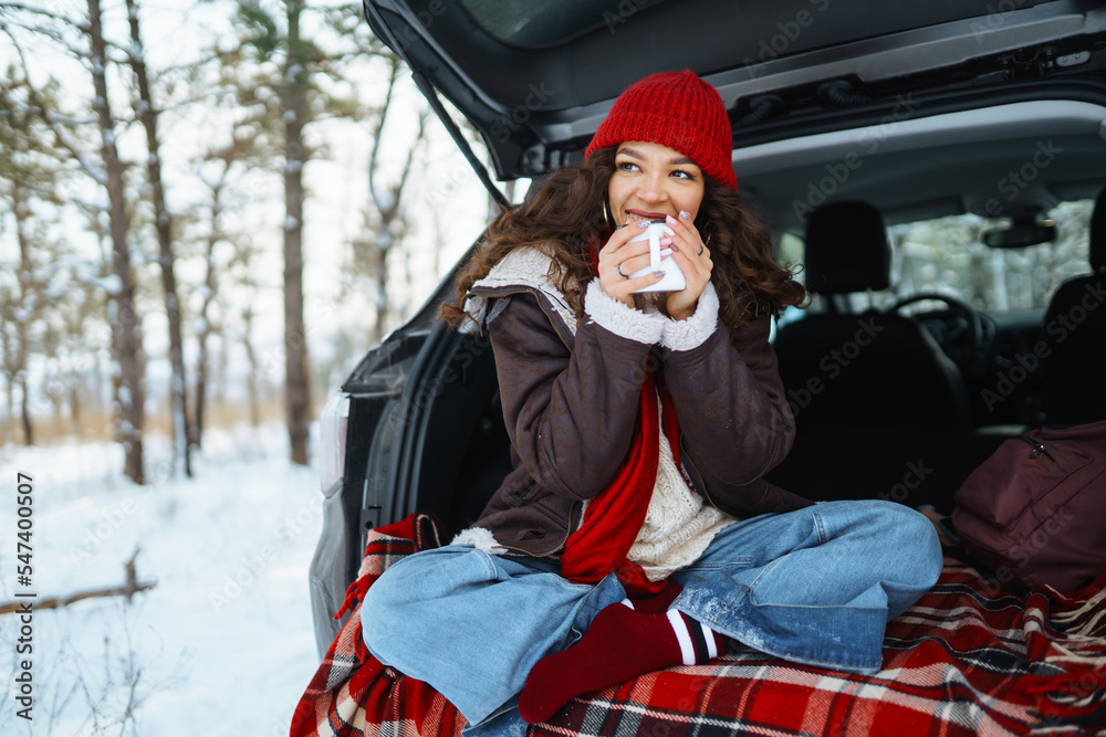 Happy woman holds a thermos and drinks tea sitting in car trunk in  winter forest. Rest, relaxation, travel, lifestyle concept.