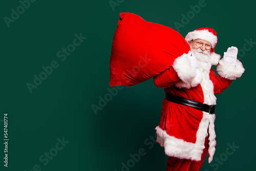 Cheerful nice peaceful Santa in eyeglasses wishes you merry Christmas waving hi hello holding keeping delivering huge sack with toys isolated over bright vivid red background photo